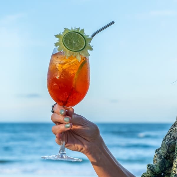 A woman holds a bright orange cocktail with a metal straw and lime slice against a backdrop of a sunny blue ocean at a Bahia beach.