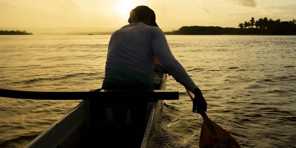 A silhouetted person paddles a Hawaiian canoe on the calm waters of the Rio das Contas during a golden sunset.