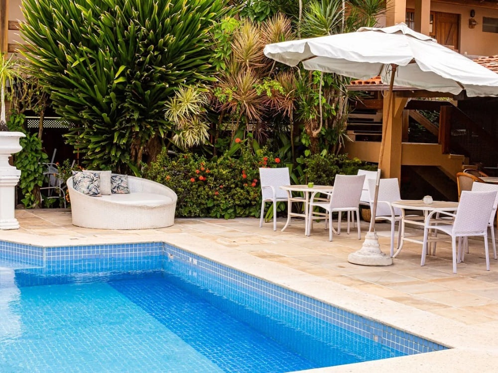 A bright blue swimming pool is surrounded by a patio with white tables, chairs, and a round lounge bed in a tropical hotel garden.