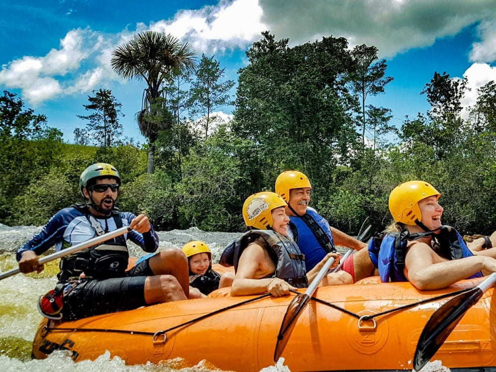 Four people wearing helmets and life jackets smiling while navigating river rapids in an orange raft.