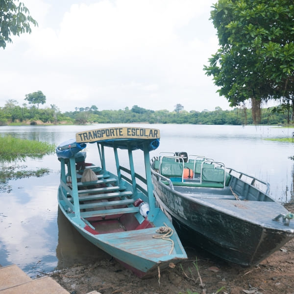 Two wooden boats docked on the riverbank, one featuring a sign reading Transporte Escolar.