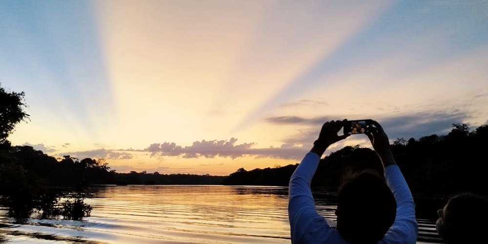 Tourist taking a smartphone photo of a golden sunrise with prominent sunbeams over the Amazon river.