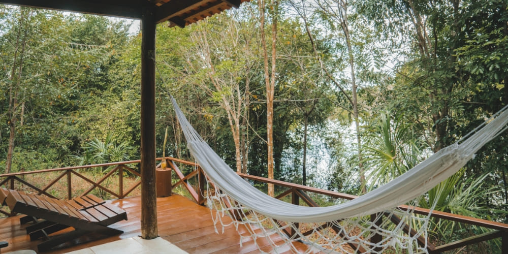 White hammock set up on the wooden balcony of a bungalow with a relaxing view of the trees and lake.