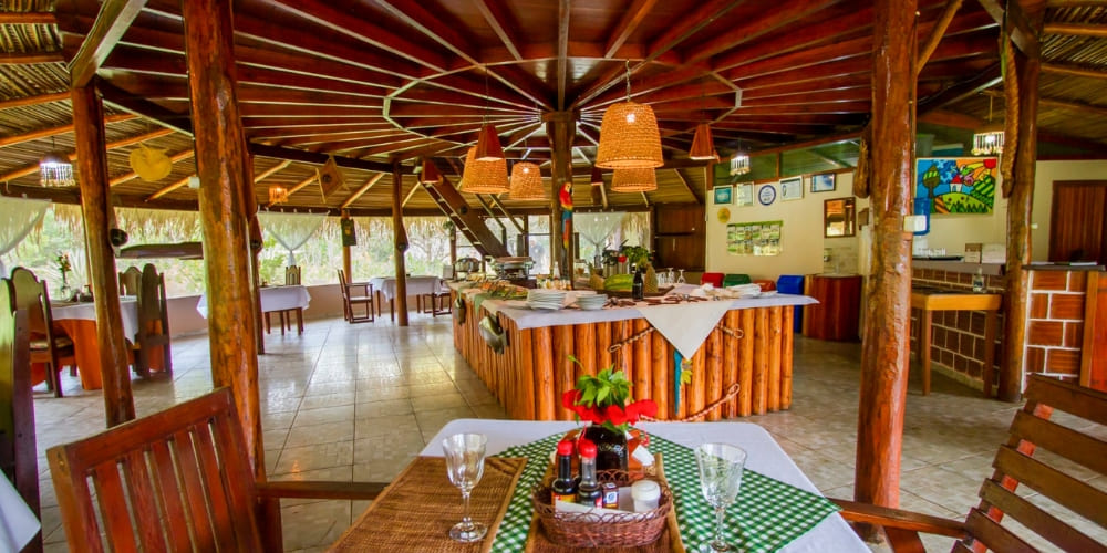 Large rustic wooden restaurant with tables and a central buffet station under a grand thatched roof.