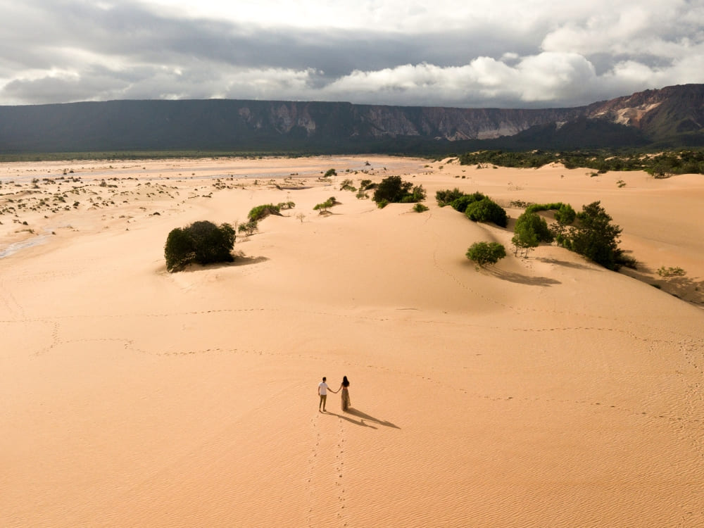 A couple holds hands while walking across the expansive golden sand dunes of the Jalapão State Park, with a mountain ridge in the background.