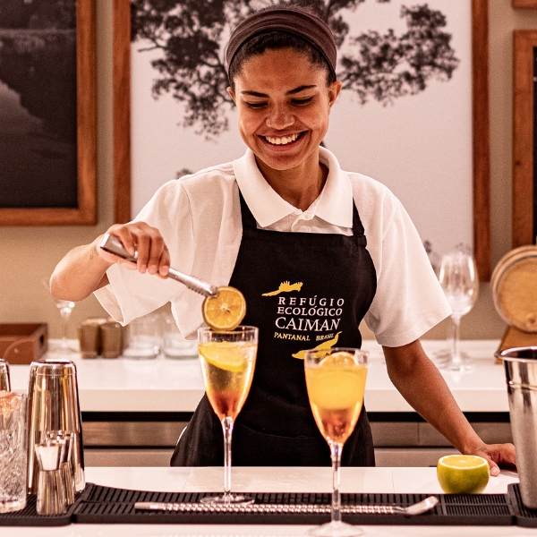 A smiling bartender wearing a Caiman apron prepares refreshing cocktails with citrus garnishes.