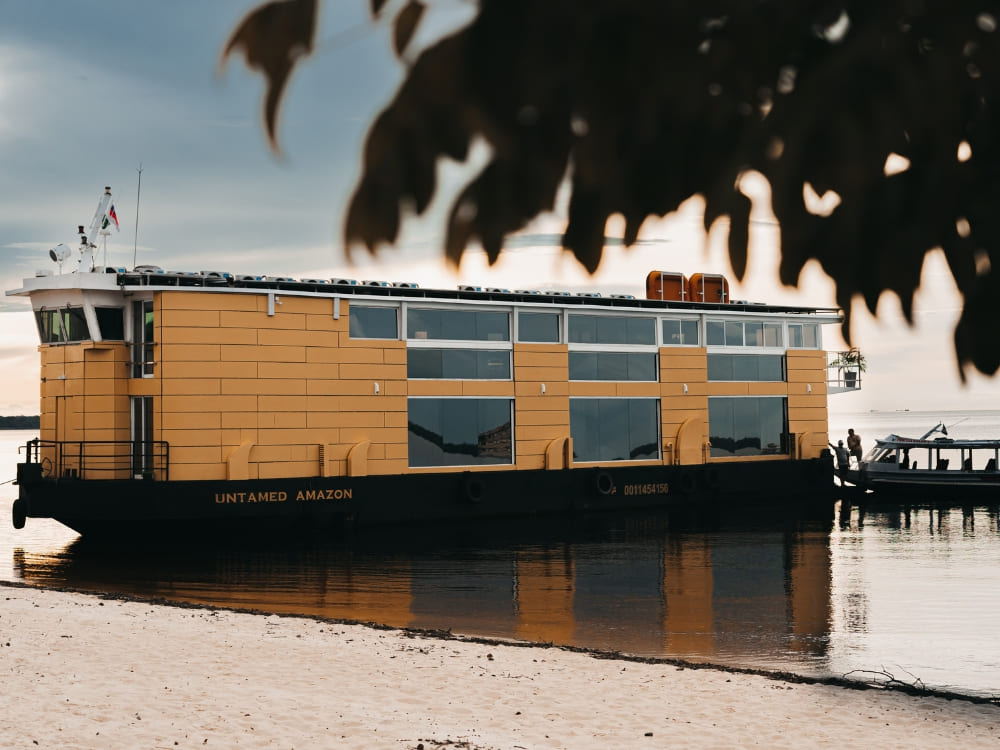 The sophisticated 3-floor vessel docked near a white-sand river beach with a smaller excursion boat alongside.