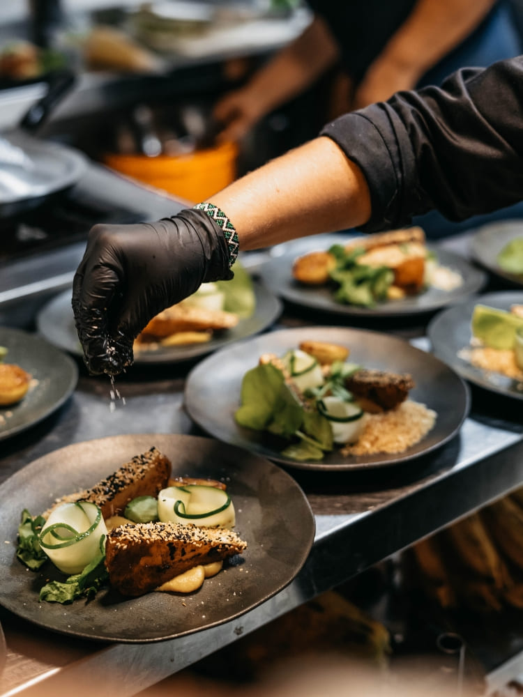 A chef wearing black gloves prepares multiple plates of gourmet food for lunch on board.