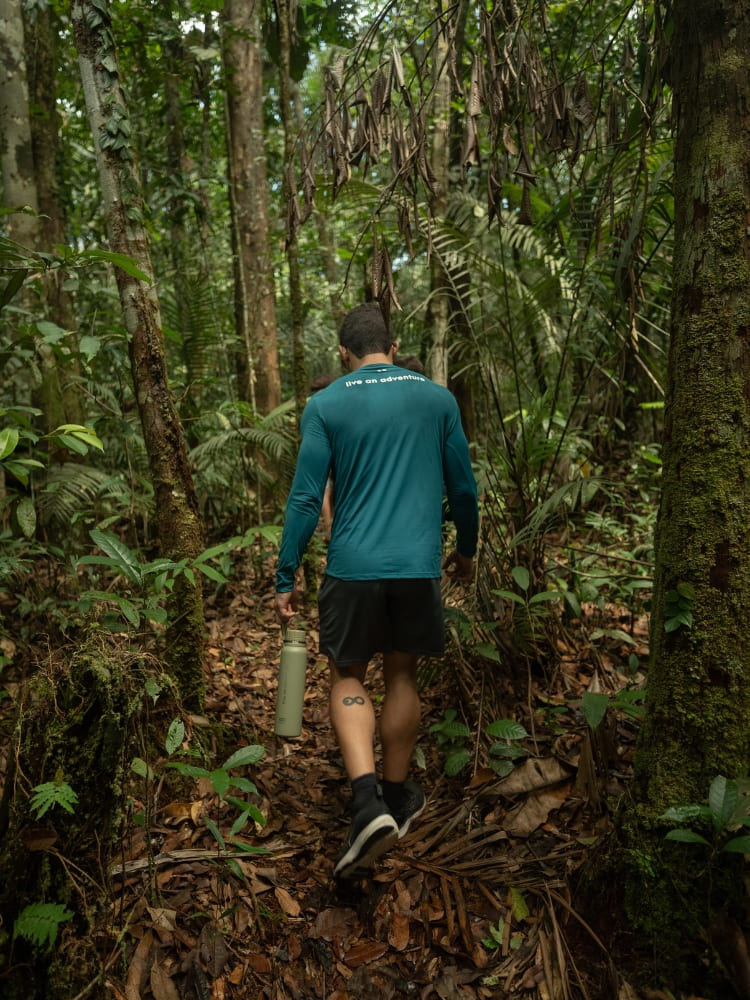 A man wearing a teal long-sleeve shirt and dark shorts walks down a leaf-covered dirt trail through a dense, green tropical forest carrying a water bottle.