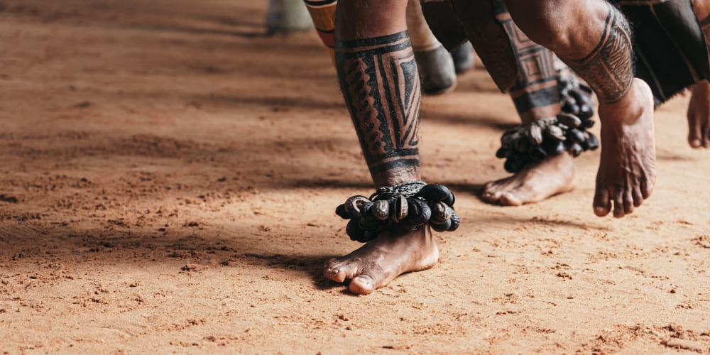  Close-up of indigenous community members wearing traditional dark ankle rattles while dancing on red dirt.