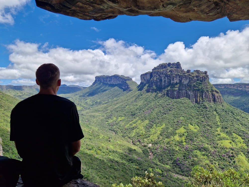 Um homem sentado sob uma saliência rochosa observando um vasto vale verde e montanhas de topo plano na Chapada Diamantina.