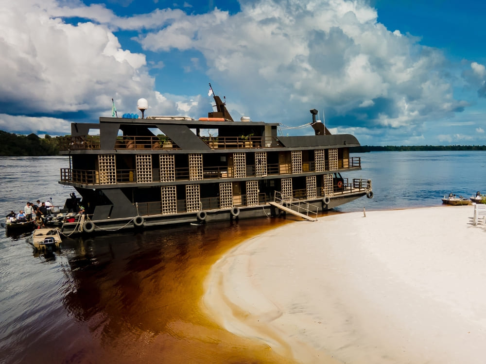 Multi-level Amazon river cruise boat docked at a sandy white beach along the dark river waters. 