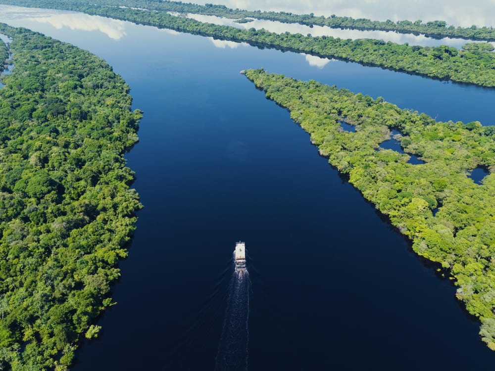 A multi-level riverboat travels along a wide, calm dark river bordered by dense green rainforest islands reflecting the sky.