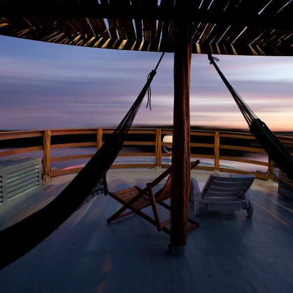 A relaxing wooden deck on an Amazon riverboat features strung hammocks and lounge chairs beneath a slatted roof during a purple and orange sunset.