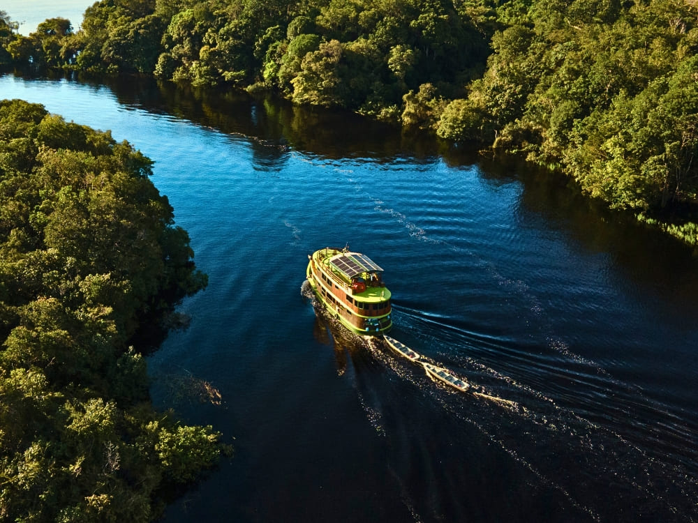 A green ecotourism riverboat with solar panels cruises down a dark river surrounded by lush Amazon rainforest.