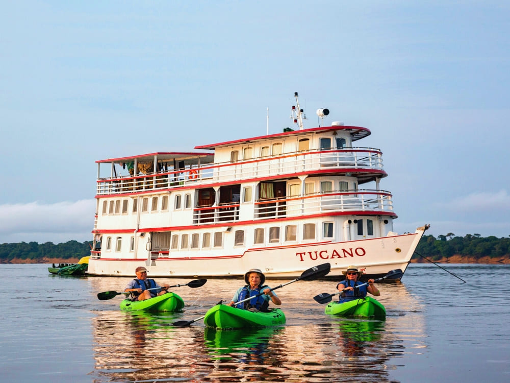 Três pessoas remam caiaques verdes em um rio calmo em frente a um barco de vários andares chamado Tucano, com árvores exuberantes na margem ao fundo.