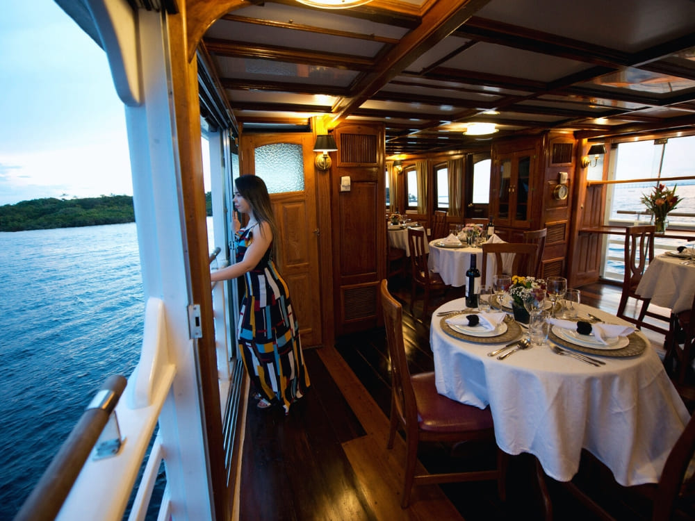 Uma mulher com um vestido colorido listrado olha pela janela da sala de jantar de um barco de madeira, que possui mesas com toalhas brancas e taças de vinho.