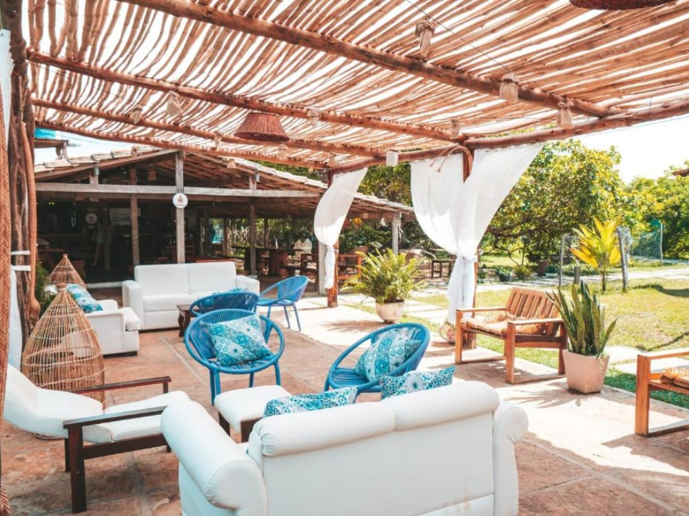 Rustic outdoor social area of an inn, covered by an irregular wooden structure and furnished with comfortable white sofas and blue chairs.