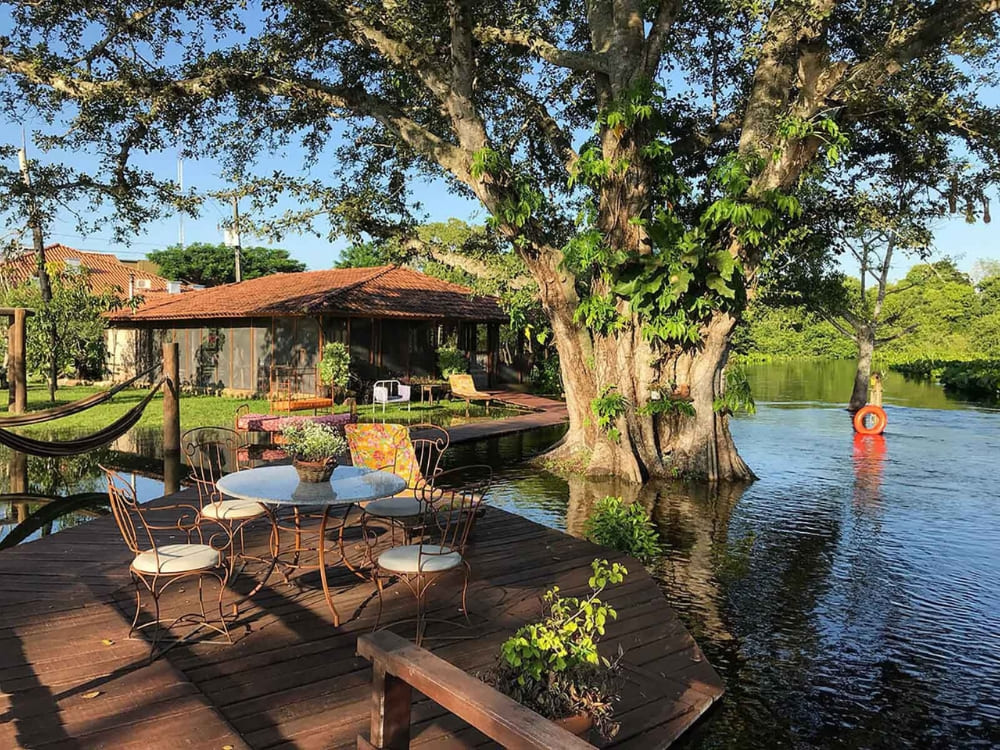 A wooden deck with a table and chairs sits beside a tranquil lake and a massive tree at Refúgio da Ilha ecolodge. 