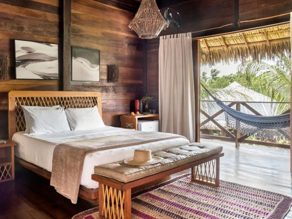 Cozy room lined with darkened wood panels at Charme Atins, with a double bed and open doors to a balcony with a hammock.