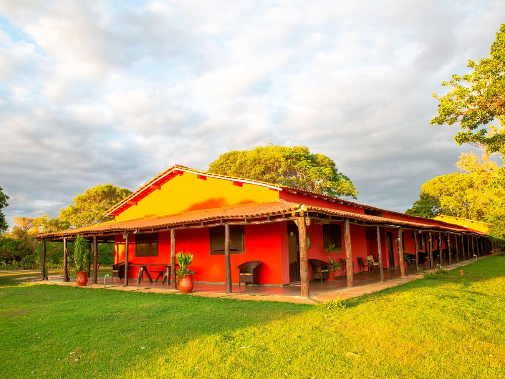 The red exterior of the SouthWild Pantanal Lodge features a shaded wooden porch for guests seeking an authentic stay. 
