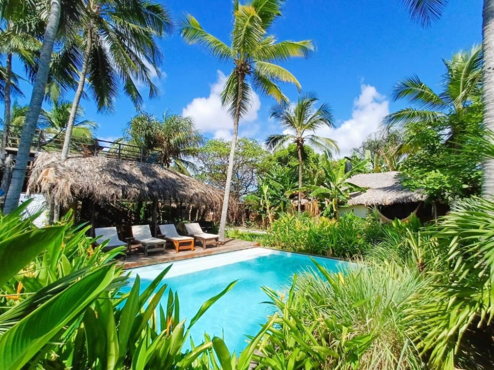 Relaxing area at Vila do Junco with a blue swimming pool surrounded by wooden sun loungers, coconut trees, and thatched kiosks.