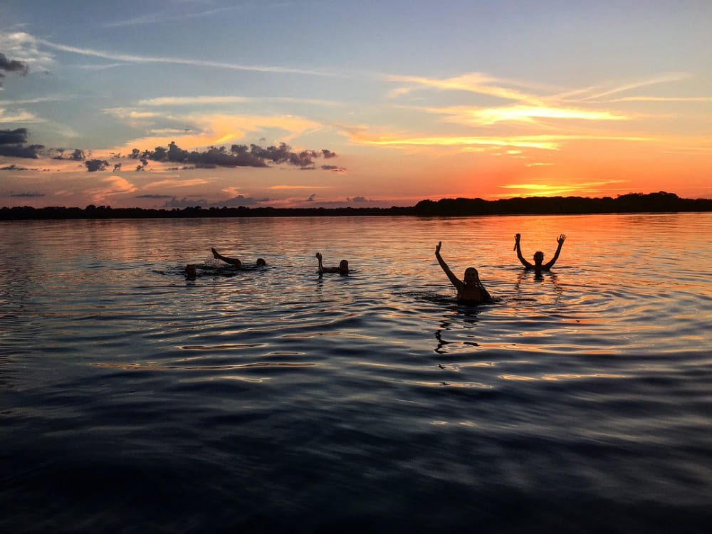 A group of travelers swims in a calm black water Amazon river during a vibrant orange sunset.