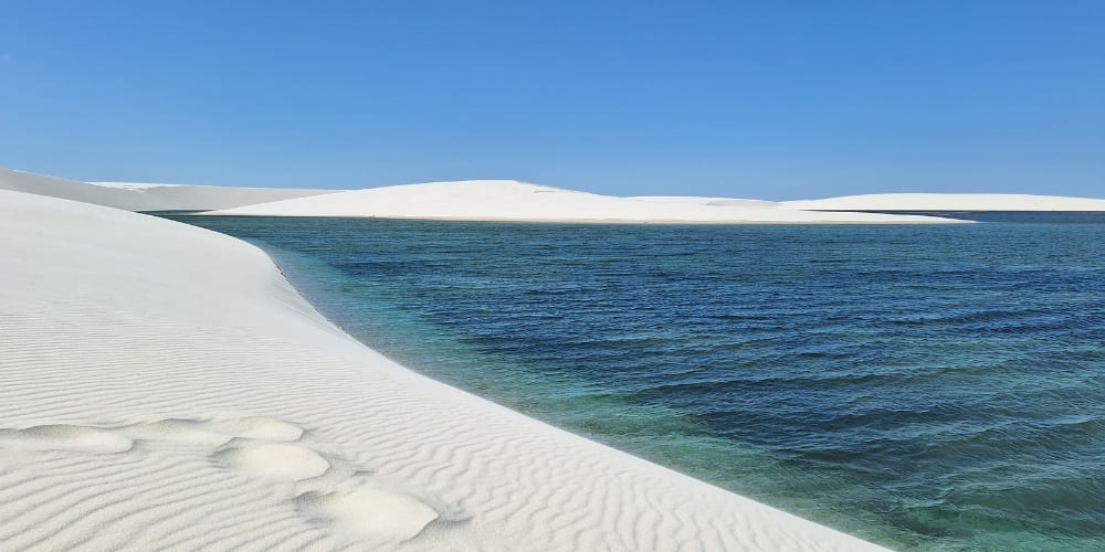 Clear blue rainwater lagoon surrounded by vast white sand dunes in Lençóis Maranhenses National Park under a bright sky.