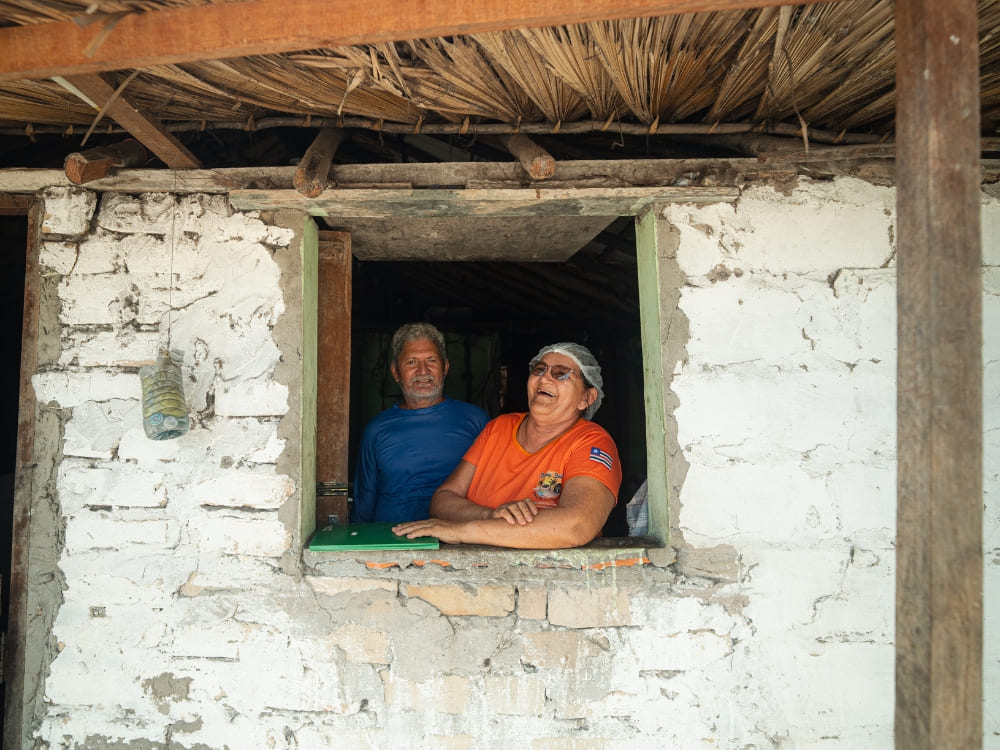 Un homme et une femme plus âgés et souriants regardent par la fenêtre d'une maison rustique blanchie à la chaux avec un toit en feuilles de palmier aux Lençóis Maranhenses.