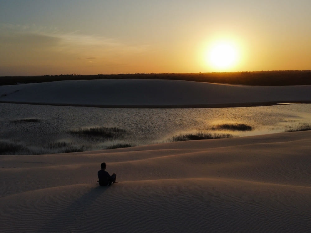 Silhouette of a person sitting on a sand dune, watching a golden sunset over the lagoons of Lençóis Maranhenses.