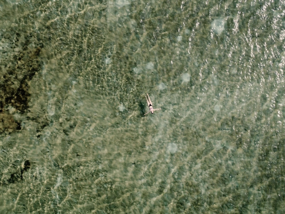 Top-down view of a person swimming in a crystal-clear rainwater lagoon in Lençóis Maranhenses, revealing the sandy bottom.
