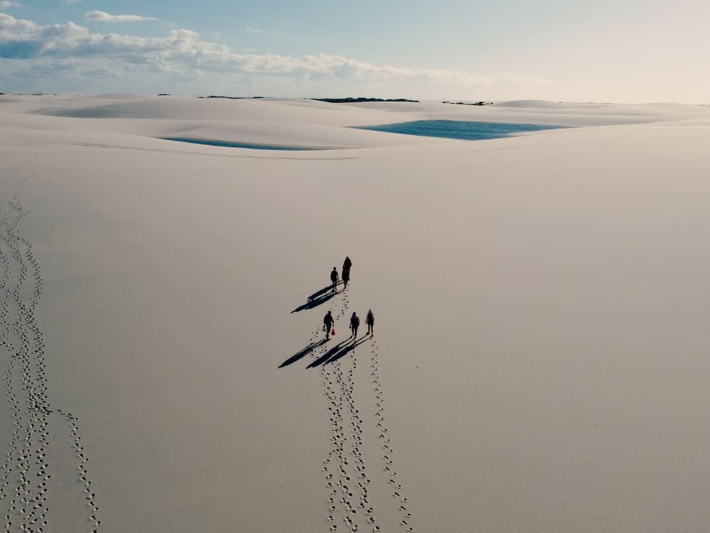 A small group of hikers casting long shadows while walking across a vast expanse of white sand dunes on a multi-day Lençóis Maranhenses trek. 