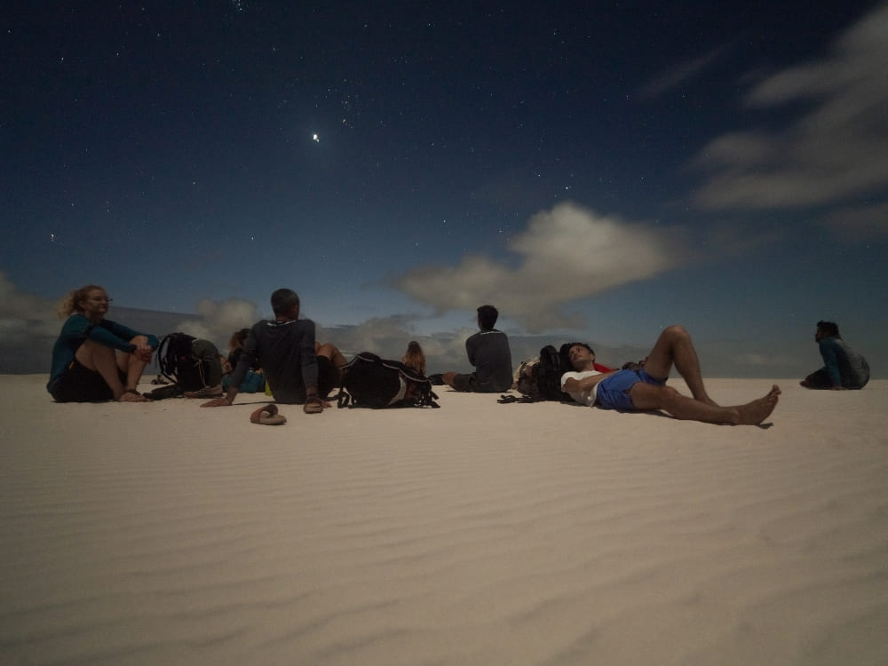 A group of travelers resting on the sand dunes under a starry night sky after a full day exploring Lençóis Maranhenses. 