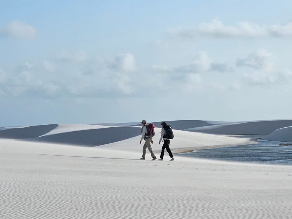 Dois mochileiros caminham pelas vastas dunas de areia branca ao lado de uma lagoa de água doce no Parque Nacional dos Lençóis Maranhenses.