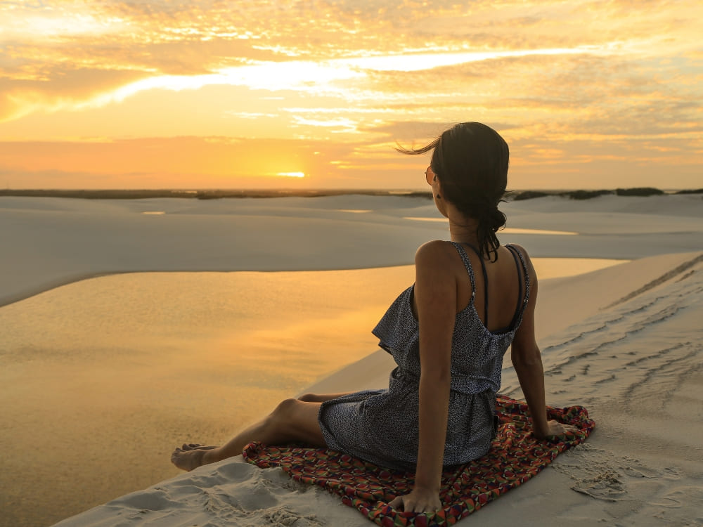 Mulher sentada em uma canga na areia branca, admirando o pôr do sol dourado refletindo nas águas das lagoas dos Lençóis Maranhenses.

