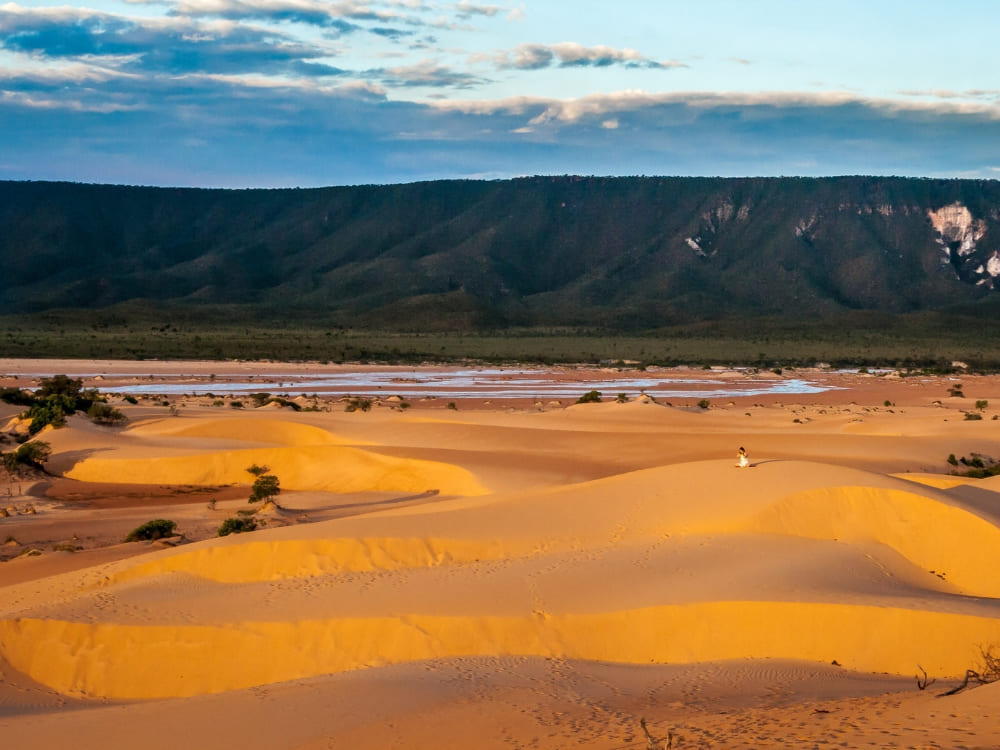A traveler sits on the golden sand dunes of Jalapão watching the sunset over the mountains.