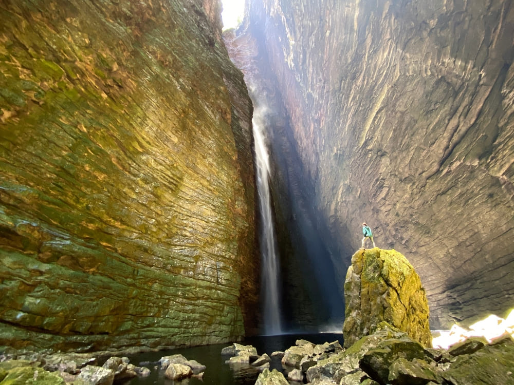 A person stands on a green mossy rock in a narrow canyon, looking at a powerful waterfall crashing into the dark water below.