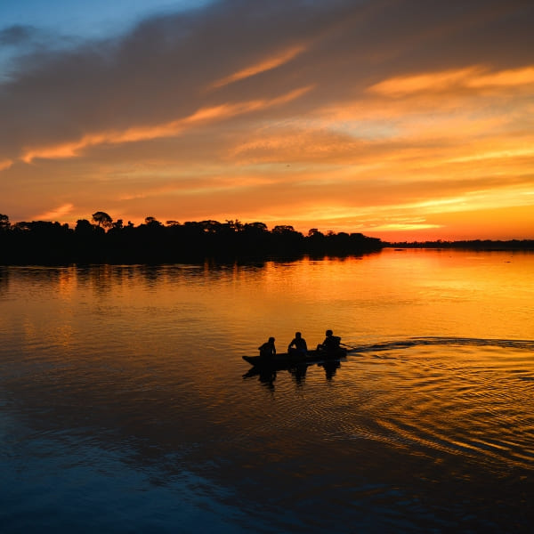 Silhueta de três pessoas em uma canoa flutuando no rio Amazonas sob um pôr do sol laranja vibrante.