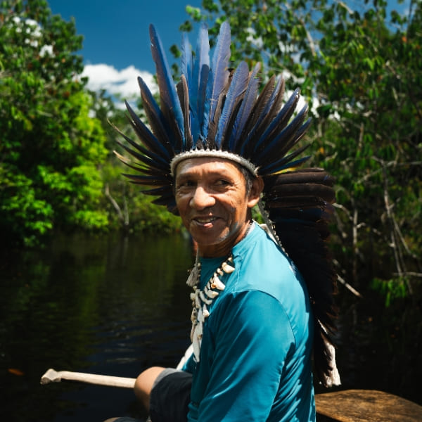Um homem indígena sorridente usando um cocar de penas azuis e um colar tradicional remando uma canoa.