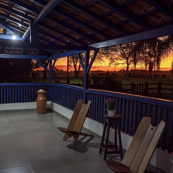 Une véranda aux balustrades bleues avec des chaises en bois donnant sur un paysage rural lors d'un coucher de soleil orange intense.