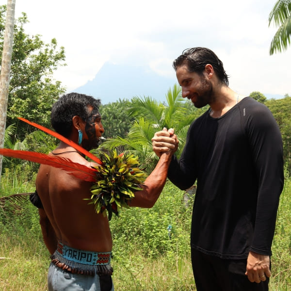 Um turista e um indígena Yanomami usando penas vermelhas tradicionais compartilhando um aperto de mão amigável na selva.
