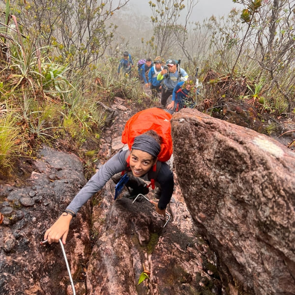 Um turista sorridente usando uma corda para subir uma trilha íngreme e rochosa durante uma expedição de trekking na Amazônia.