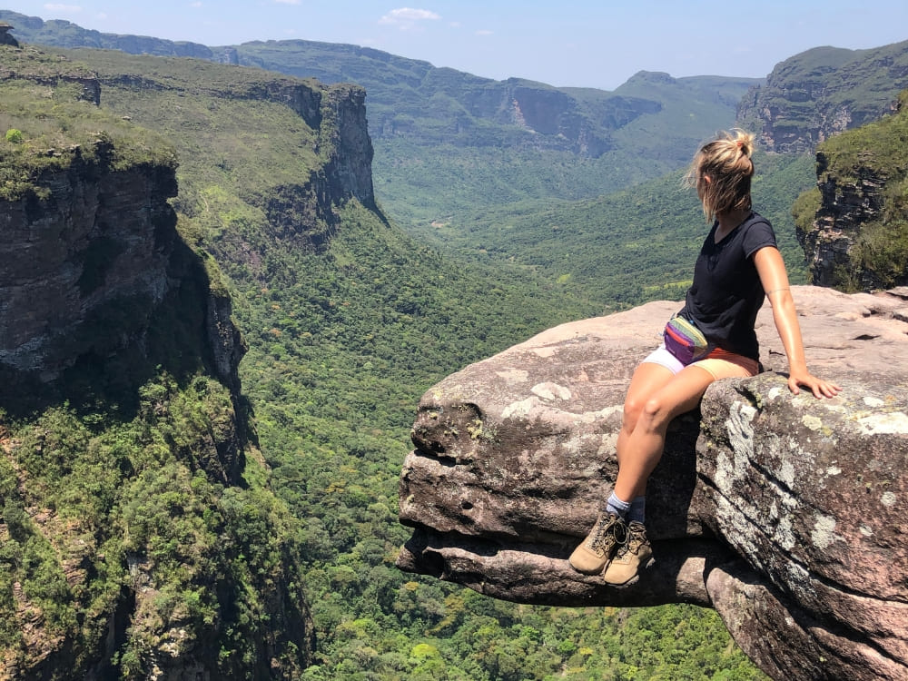 Woman hiker resting on a rocky cliff edge, overlooking a vast, lush green canyon and distant plateaus during an ecotourism adventure.
