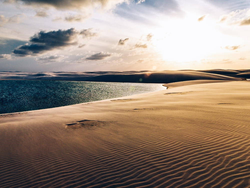 A calm blue lagoon sits beside vast, textured sand dunes under a partly cloudy sky, showing the best time to visit Lençóis Maranhenses.