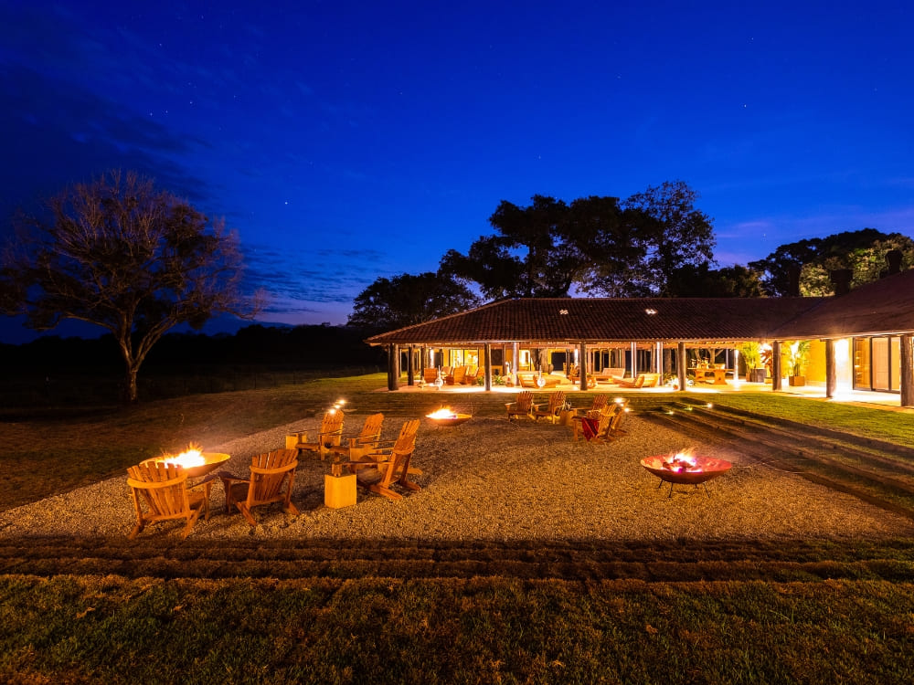 The exterior lounge of Casa Caiman at night, featuring fire pits and wooden chairs under a deep blue starry sky.