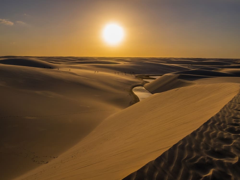 De zon gaat helder onder over glooiende zandduinen en een ondiep stroompje dat door het Lençóis Maranhenses national park in Brazilië kronkelt.