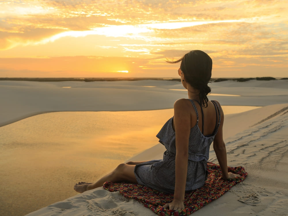 Een vrouw zit op een handdoek bovenop een duin, genietend van het warme weer in Lençóis Maranhenses, en kijkt naar de zonsondergang over een serene lagune.
