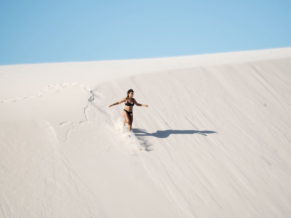 Een vrouw in een zwarte bikinitop en korte broek rent vrolijk van een steile witte zandduin af tijdens het verkennen van Lençóis Maranhenses.