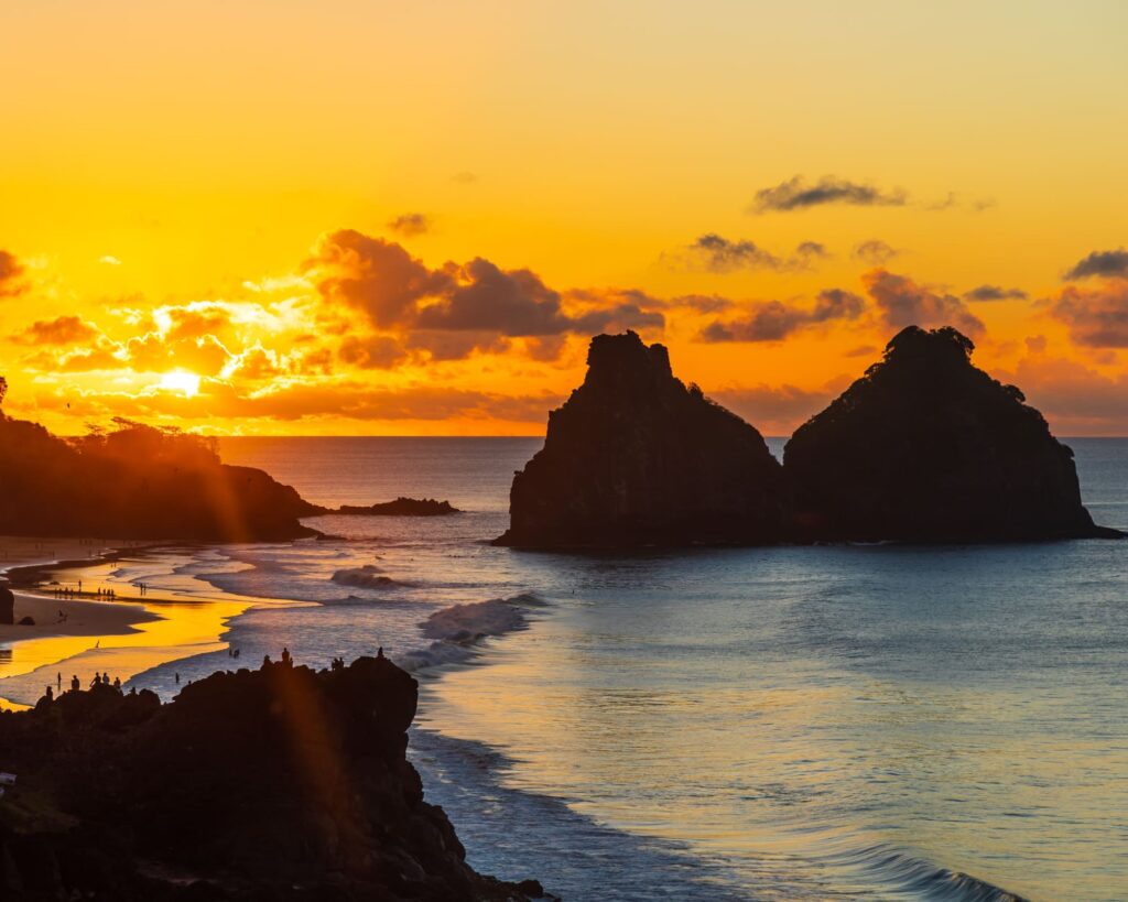 Golden sunset over the Dois Irmãos rock formation at a Fernando de Noronha beach, with silhouettes of people watching the glowing orange sky and gentle waves.
