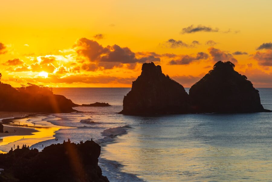 Golden sunset over the Dois Irmãos rock formation at a Fernando de Noronha beach, with silhouettes of people watching the glowing orange sky and gentle waves.